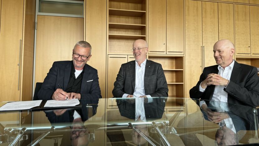 Drei Männer sitzen an einem Konferenztisch, einer unterzeichnet einen Vertrag.